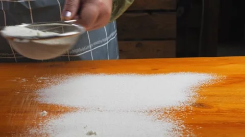 Flour is slowly poured evenly onto a light wooden table from a sieve in a w.. Stock Footage 268742822