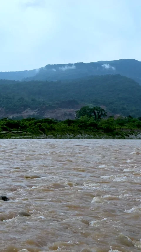 Flowing river with muddy waters and lush green hills under a cloudy sky in .. Stock Footage 338414414