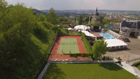 Flying back over players playing tennis on a court in green city park with pool Stock Footage 212945395