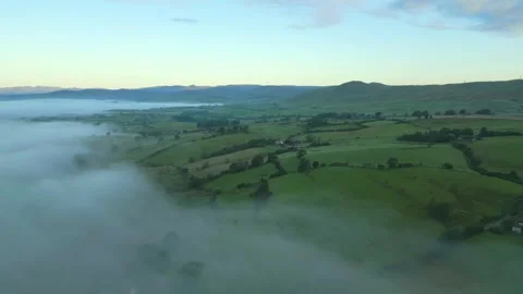 Flying high over mist towards green patchwork fields of rural England at Stock Footage 248495023