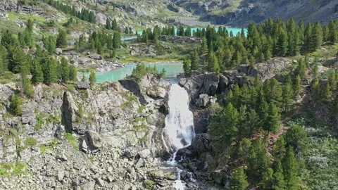 Flying over a small waterfall over a cliff with a forest and a turquoise lake on Stock Footage 264510914