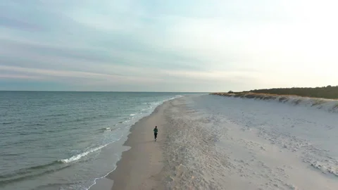 Flying over wide white beach with female jogger running along the shoreline Stock Footage 153265624