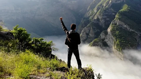 Flying over young man standing victoriously at the edge of the cliff at sunrise Stock Footage 90574870