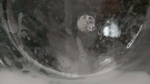 Foam bubbles and dark clothes swirling behind the wet glass door of a running 動画素材 348094556