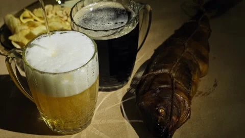 Foaming dark and light beer poured in mugs and fresh smoked fish, close-up. Видео 259142615