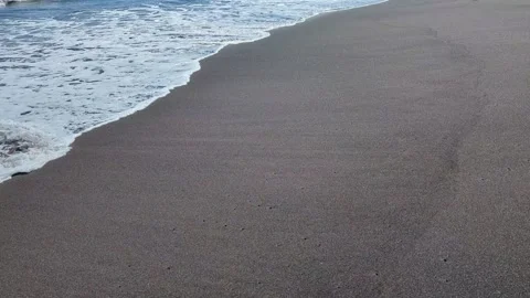 Foaming ocean water gently washes over dark volcanic sand. The waves recede.. Video stock 331179544
