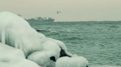 Foaming wave is breaking on the ice coverd stone. Cargo container ship sailing. Video stock 55425354