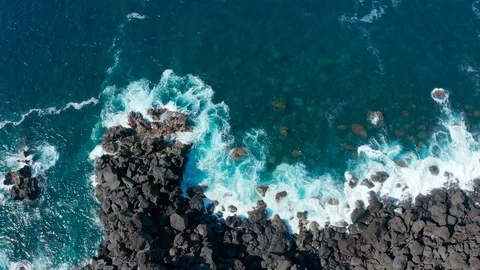 Foaming waves roll on big black stones on the shore of blue ocean. Aerial slow Stock Footage 115098684