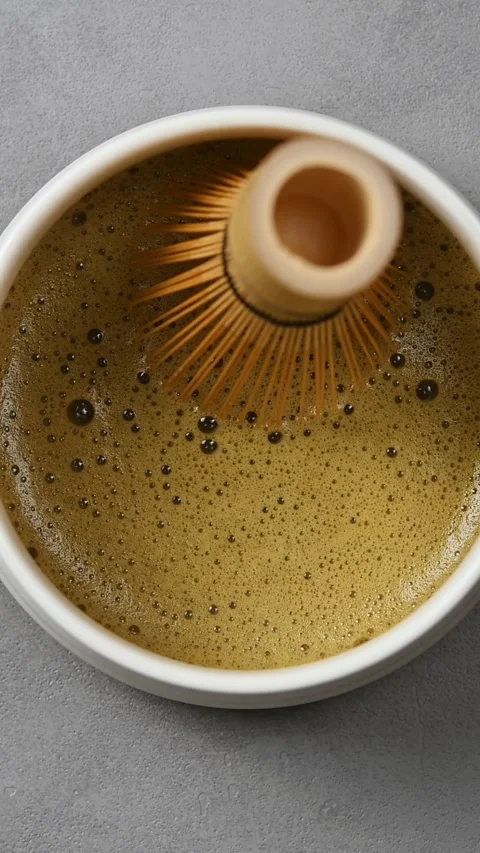 Foamy hojicha tea texture is shown in ceramic bowl as bamboo whisk moves across Stock Footage 343495331