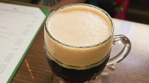 Foamy mug of dark lager resting on a wooden tavern table, showing thick creamy Stock Footage 339234948