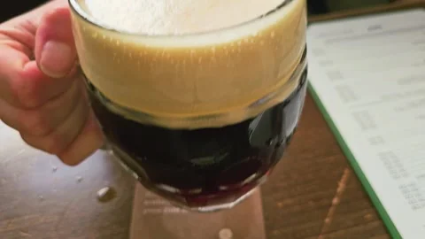 Foamy pint of dark beer in a glass mug held by a hand at a pub table, showing Stock Footage 339237448