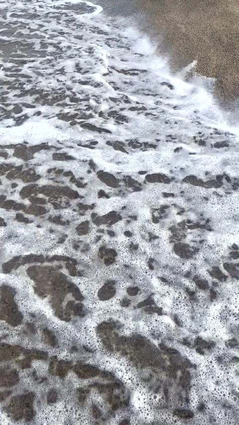 Foamy sea wave gently washes over sandy beach, creating natural white sea foam Видео 338074651