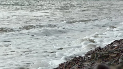 Foamy sea waves roll onto a sandy beach under an overcast sky, creating a calm Stock Footage 323980941