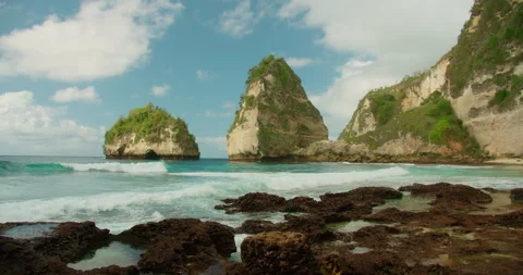 Foamy surf rushing over reef rocks toward a towering mountain peak by the shore Vídeo Stock 344778624
