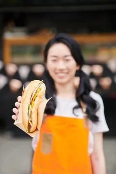 Focus on appetizing hotdog in paper held by young Asian female vendor Stock Photos