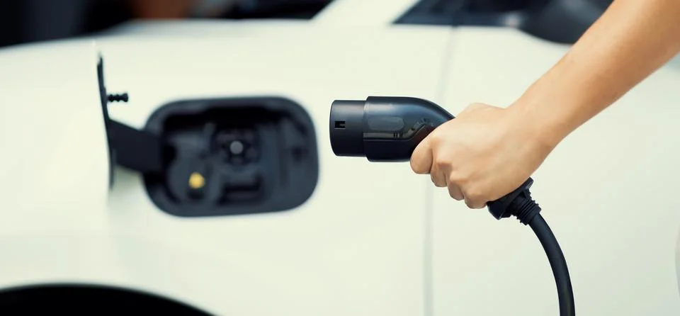 Focus hand hold EV plug with blur image of progressive car at charging station. Stock Photos