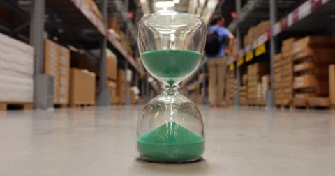 Focus on hourglass measuring time in large warehouse aisle, man pass by Stock Footage 319014678