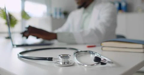 Focus on stethoscope lying on table and African-American doctor typing on laptop Stock Photos