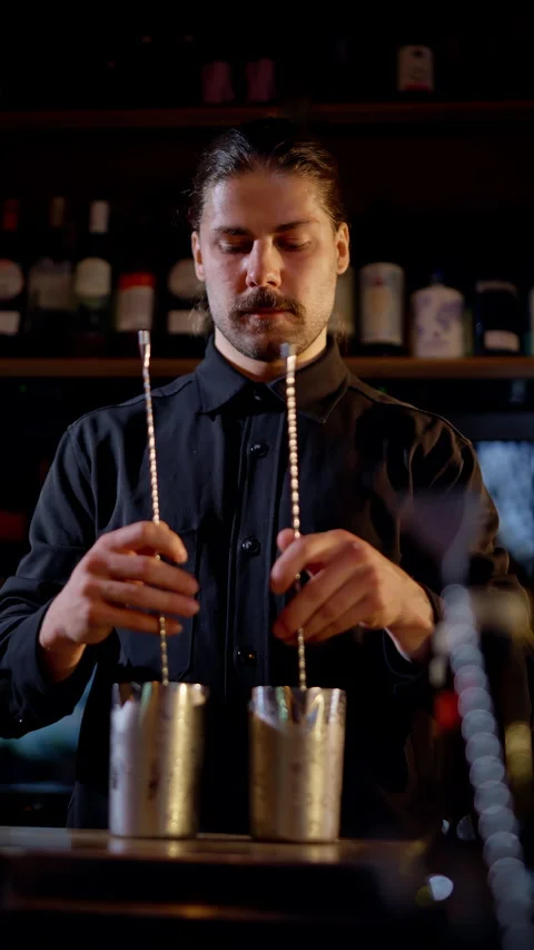 Focused bartender in a dimly lit bar expertly stirring two stainless mixing tins Stock Footage 318540149