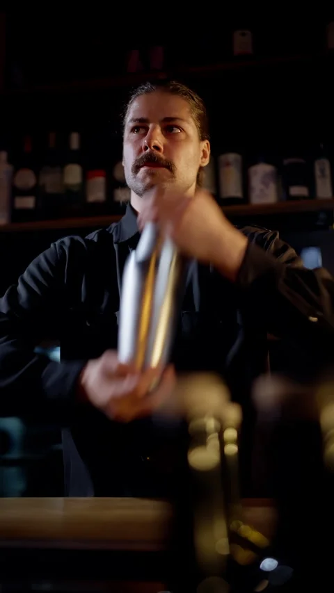 A focused bartender in a dimly lit lounge shaking a stainless cocktail shaker Video stock 318538788