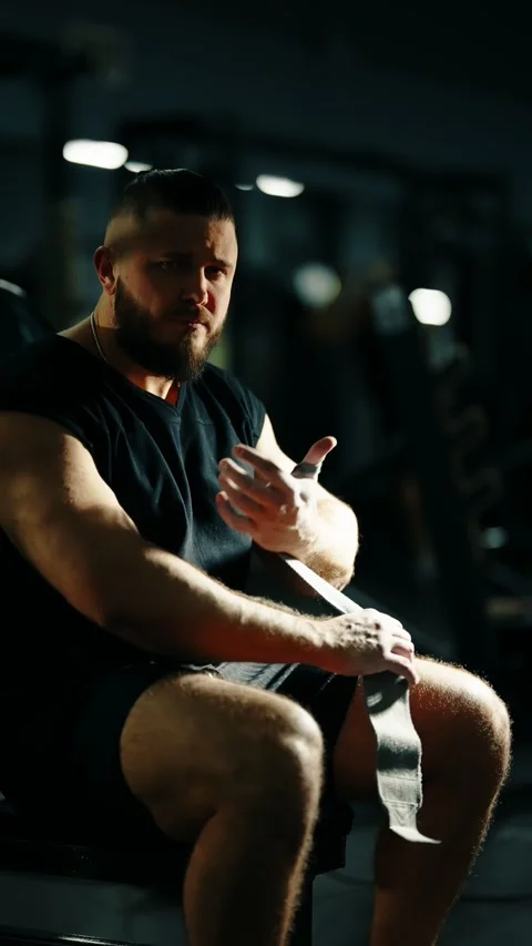 Focused bodybuilder seated on a gym bench wrapping lifting straps around his Vidéo 329556086