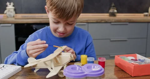 Focused boy applies paint with brush onto airplane wings at desk Stock Footage 323974504