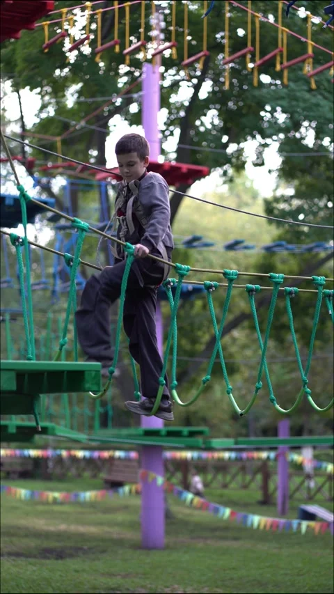A focused boy in a climbing harness balances on a swinging green rope bridge Stock Footage 345285284