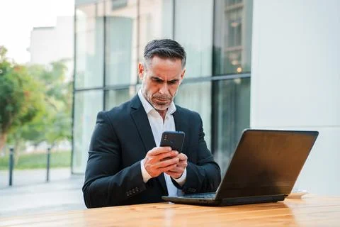Focused businessman using smartphone while working on laptop outdoors in a .. Stock Photos