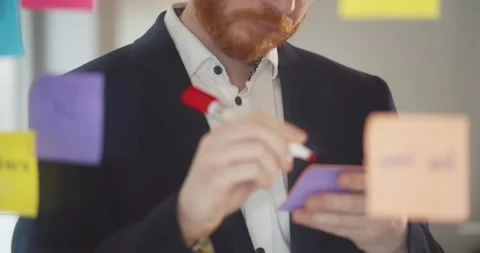 Focused businessman writing on sticky notes and attaching them to glass wall Video stock 148231372
