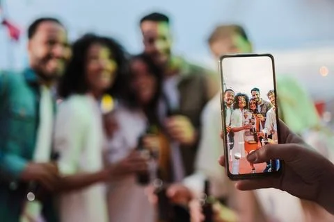 Focused on the cellphone screen. Blurred group of a young adult friends shooting Stock Photos