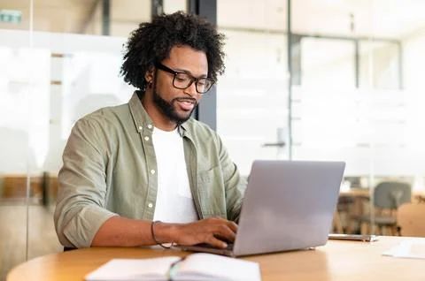Focused curly handsome man using laptop computer in contemporary coworking space Stock Photos