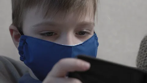 Focused cute boy in protective mask using smartphone sit at home sofa during the Video stock 129380568