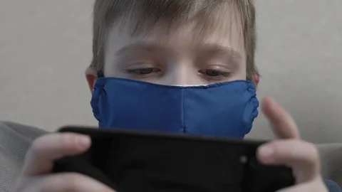 Focused cute boy in protective mask using smartphone sit at home sofa during the Video stock 129380601