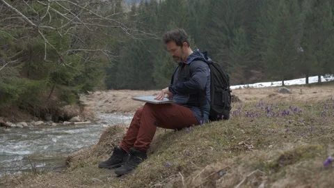 Focused digital nomad working on a laptop by a tranquil mountain river surr.. Vídeo Stock 331207560