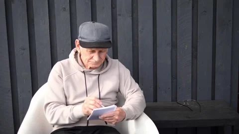 Focused elderly person writing notes in notebook, sitting on white chair near Stock Footage 306924357