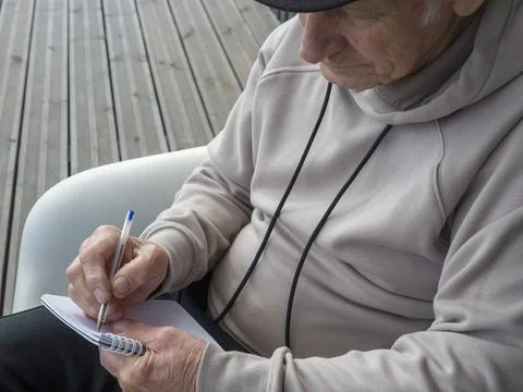 Focused elderly person writing notes in notebook, sitting on white chair near Stock Photos