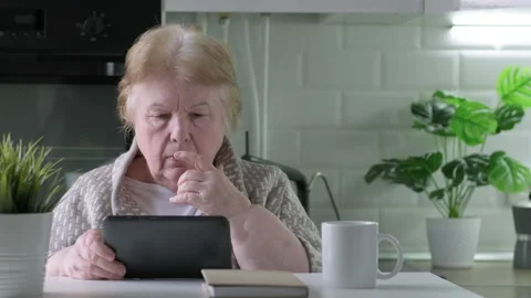 Focused elderly woman sit at table at home kitchen browse internet on pad gadget Stock Footage 195212061