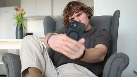 With a focused expression, the guy sits in a chair and records something on his  Stock Footage 302633789