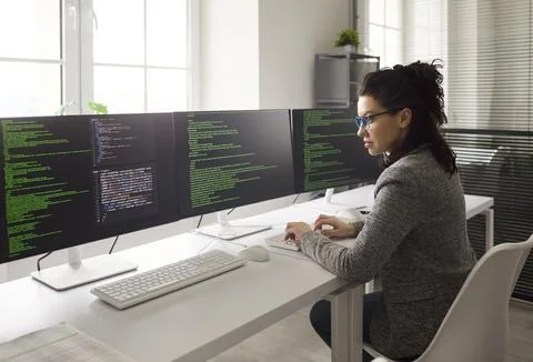 Focused female developer coding software on multi monitor workstation in office Stock Photos