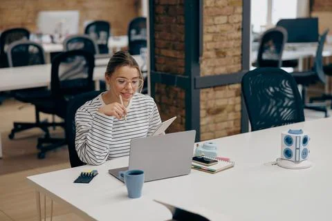 Focused female freelancer is making notes while use laptop sitting in modern Stock Photos