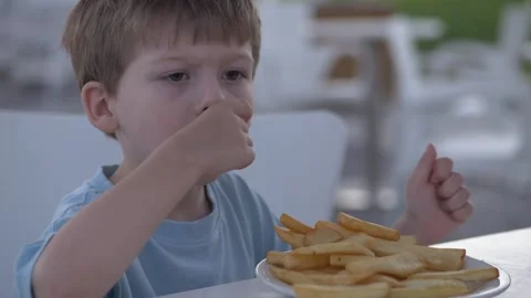 A focused four-year-old boy picks up fries with his fingers, enjoying the snack Stock Footage 317317177