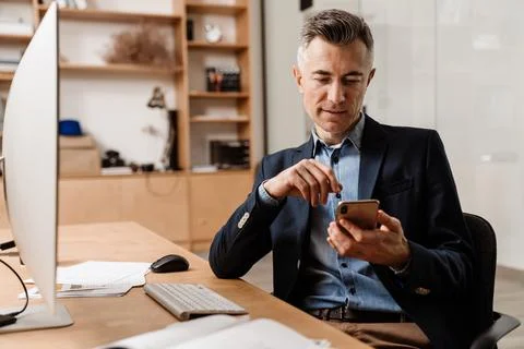 Focused grey man using mobile phone while working with computer Foto stock