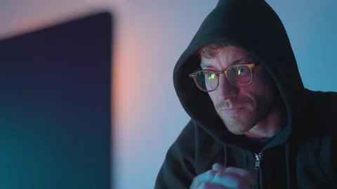 Focused hacker in glasses and dark hoodie sitting in front of a computer screen Stock Footage 332445288