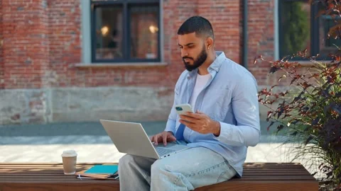 Focused indian man revising information between laptop and smartphone outdoors Stock Footage 250075009