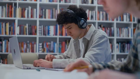 Focused Male Duo at a Library Table: Laptop, Headphones, and Paper Notes 스톡 동영상 335124742