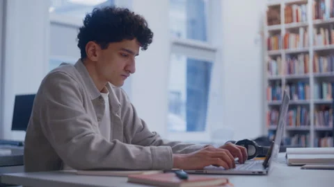 Focused Male Student Working on Computer at Desk Surrounded by Books 스톡 동영상 335123961