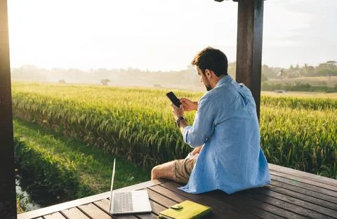 Focused man browsing smartphone while working on laptop outdoors Stock Photos