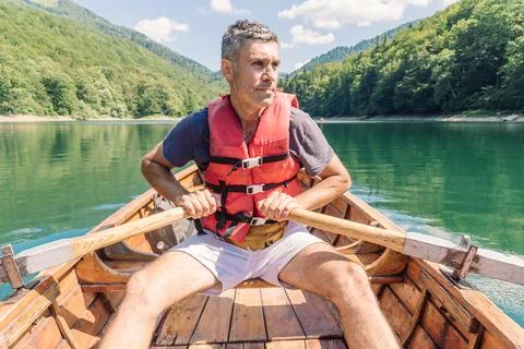 Focused man grips both oars rowing wooden boat on mountain lake in life jacke Photos