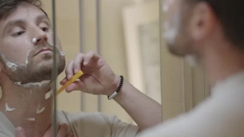 Focused man looking in the bathroom mirror while shaving with foam and a Stock Footage 335201176