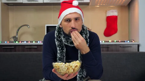 Focused man in New Year red Santa Claus hat eating popcorn, watching TV Stock Footage 136660952